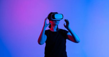 Bir kadın VR kulaklığını ayarlamak için uzanıyor, parlak mavi ve pembe neon ışığı etrafını sararken, büyüleyici bir seansa hazırlanıyor..