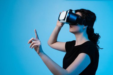 VR kulaklık takan bir kadının yan profili ve mavi arkaplandaki sanal arayüzle etkileşime geçmek için öne uzanıyor.