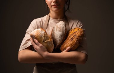 Bir kadın fırıncı, çeşitli kabuk dokuları ve un desenleriyle sıcak stüdyo ışıklarıyla sunulan üç adet rustik el yapımı ekmek somunu tutar..