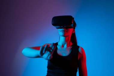 VR kulaklık takan bir kadın canlı neon ışıklarda sanal elementlerle etkileşime girer ve elini dijital ortamda hareket ettirir.