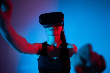 VR kulaklık takan bir kadın, neon ışıklı bir ortamda sanal kontrolle etkileşime geçiyor ve fütürist ve büyüleyici bir sanal gerçeklik deneyimi yaratıyor..