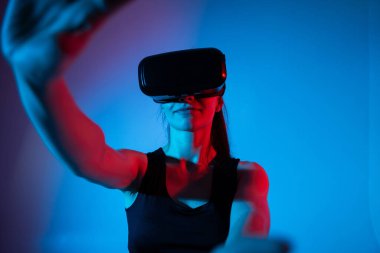 Kadın kullanıcı sanal gerçeklikle bütünüyle meşgul, kollarını ileri hareket ettirirken neon ışıklarının altındaki dijital bir alanla etkileşime geçiyor.