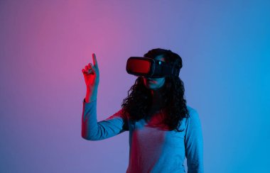 VR kulaklık takan genç bir kadın, mavi ve kırmızı neon ışık altında sanal arayüzle etkileşime giriyor, stüdyoda dijital ortamları keşfediyor..