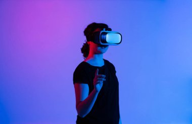VR kulaklık takan kadın, koyu mavi ve pembe neon ışıklarla çevrili sanal elementlerle etkileşime geçmek için parmağını kaldırır..