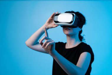 Profildeki kadın, VR kulaklığını ayarlıyor ve sanal içeriğe yönelik jestler yapıyor, mavi arka planda dalgın dijital ortamı keşfediyor