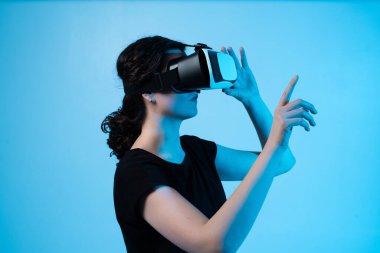 Sanal kontrolleri ve hareket arayüzünü keşfeden, mavi arkaplandaki dijital ortamla etkileşime geçmek için VR kulaklık takan bir kadın