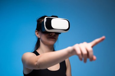 VR kulaklık takan genç bir kadın mavi arka planda sanal gerçeklik teknolojisi ve dijital ortamları keşfederek sanal arayüzle etkileşime geçiyor..