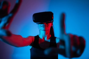 Genç bir kadın, bir VR kulaklık kullanarak iki elini de ileri uzatarak neon ışıkta etkileşimli bir sanal arayüze bağlanıyor..