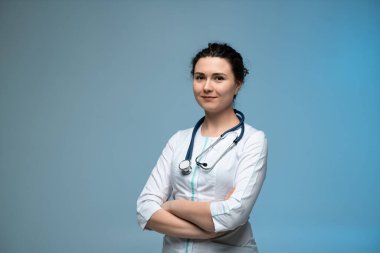 Steteskoplu, kendine güvenen, beyaz üniformalı kadın doktor, mavi arka planda poz veriyor, modern profesyonel sağlık hizmetlerini temsil ediyor.