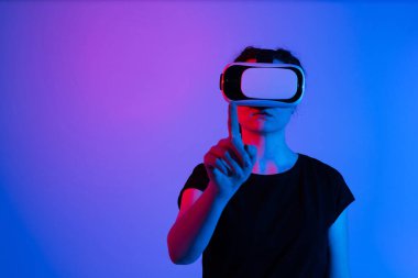 Sanal dünyayla etkileşim kurmak için VR kulaklık kullanan ve elini kaldıran bir kadın. Minimalist kompozisyon ve neon ışıklandırma ile donanımlı dijital teknoloji kavramı.