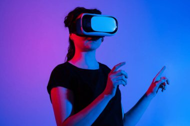 Sanal gerçeklik kulaklığı takmış bir kadın el hareketleriyle dijital arayüzle etkileşime geçiyor. Neon stüdyo ışıklandırmalı fütüristik büyüleyici teknoloji konsepti.