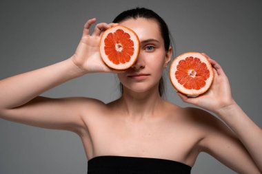 Tarafsız arka planda yüzünün yanında taze greyfurt parçaları tutan gülümseyen kadın, sağlıklı yaşam tarzı, doğal güzellik, vitaminler, sağlık konsepti.