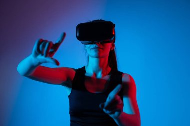 Kadın kullanıcı bir VR kulaklık takıyor ve tek eliyle öne bakıyor. Jest kontrolü kavramı, sanal etkileşim, büyüleyici teknoloji ve dijital arayüz navigasyonu.