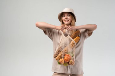 Sebze, meyve ve ekmekle dolu yeniden kullanılabilir bir alışveriş çantası tutan genç bir kadın. Sürdürülebilir market alışverişi, eko yaşam tarzı ve sağlıklı beslenme kavramı.