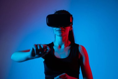 Kadın sanal gerçeklik kulaklığı takıyor ve dramatik neon ışıkları altında sanal alanı işaret ediyor. Kapsamlı dijital deneyim, kullanıcı kontrolü, siber teknoloji ve gelecekteki arayüz kavramı.