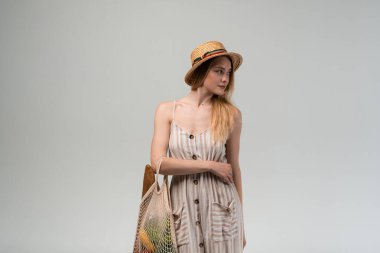 Hasır şapka takan ve yaz elbisesi giyen genç bir kadın alışveriş torbası tutuyor. Sürdürülebilir yaşam tarzını, çevre dostu alışkanlıklarını ve sağlıklı gıda alışveriş kavramlarını vurgulayan temiz bir stüdyo portresi.