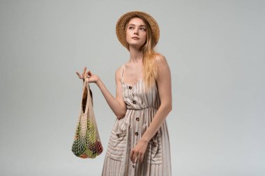 Stüdyo yaşam tarzı, elinde yeni üretilen bir alışveriş poşeti tutarken hasır şapka ve çizgili bir yaz elbisesi giyen genç bir kadının görüntüsü. Çevre alışverişi ve sürdürülebilirlik kavramı.