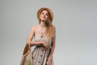 Stüdyoda içi alışveriş torbalarıyla dolu, hasır şapka ve çizgili elbiseyle poz veren gülümseyen genç bir kadın. Eko yaşam tarzı ve sağlık temaları için en temiz imaj.