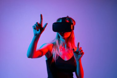 VR kulaklık takan genç bir kadın sanal arayüzü işaret ediyor ve onunla etkileşime geçiyor. Neon kırmızı ve mavi stüdyo ışıklandırmalı gelecekteki oyun konsepti.