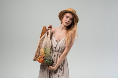 Stüdyoda hasır şapka takan ve çizgili elbise giyen genç bir kadının yeniden kullanılabilir bir alışveriş poşeti tutarken çekilmiş fotoğrafı. Sürdürülebilir yaşam tarzı, eko-alışveriş ve sağlıklı gıda konsepti.