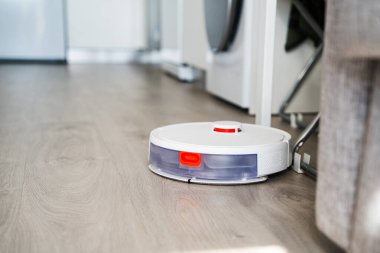 Robotik elektrikli süpürge mobilya ve aletlerin yanındaki ahşap zeminde hareket ediyor, akıllı navigasyon, akıllı ev yaşamı, zaman tasarrufu teknolojisi ve modern ev temizliği gösteriyor..