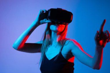 VR kulaklık takan genç bir kadın, aygıtı sanal uzayda ileri doğru uzanırken ayarlıyor, daldırıcı teknolojiyi, metatik etkileşimi ve dijital gelecek kavramlarını sembolize ediyor.