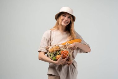 Kova şapka takan gülümseyen genç bir kadın içinde taze sebze ve ekmek dolu kullanılabilir bir market poşeti tutuyor. Eko-alışveriş kavramı, plastiksiz yaşam tarzı, sağlıklı gıda seçimleri ve sürdürülebilir günlük yaşam.