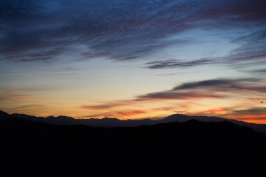 Yumuşak pastel günbatımı renkleri, karanlık dağ siluetleri üzerinde gökyüzünde solup, arka plan, seyahat, doğa ve farkındalık temaları için ideal huzurlu bir akşam manzarası yaratır..