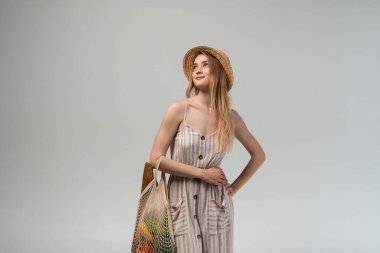 Yazlık elbise ve hasır şapkayla poz veren kendine güvenen genç bir kadın elinde içinde taze ürünler bulunan bir market poşeti tutuyor. Ekolojik yaşamı, sürdürülebilir alışverişi ve sağlıklı yaşam tarzını temsil eden en küçük stüdyo görüntüsü.