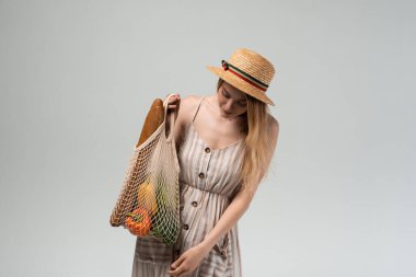 Hasır şapkalı ve açık renk yazlık elbiseli genç bir kadın, elinde ekmek ve taze ürünlerle yeniden kullanılabilir bir alışveriş poşeti tutarken aşağıya bakıyor. Çevre alışverişi ve sürdürülebilirlik hakkında stüdyo yaşam tarzı.