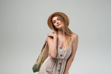 Hasır şapka ve açık yaz elbisesiyle poz veren şık genç bir kadın elinde alışveriş torbası tutarken. Temiz stüdyo görüntüsü ekolojik yaşam ve bilinçli tüketime odaklandı.