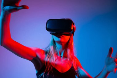 Kadın VR kullanıcısı dijital bir arayüze dokunur gibi dışarıya uzanır. Neon kırmızı ve mavi ışık büyüleyici teknoloji ve sanal gerçeklik etkileşimini temsil eden bir siber punk atmosferi yaratır..