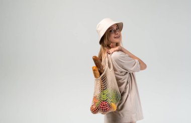 Kovalı şapka takan ve büyük beden tişört giyen genç bir kadının stüdyo portresi, üzerinde ekmek ve taze sebzelerle birlikte yeniden kullanılabilir bir alışveriş torbası taşıyor. Çevre yaşam tarzı ve sürdürülebilir alışveriş hakkında en az imaj.