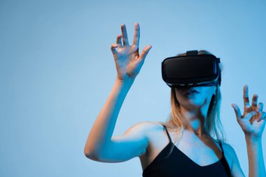 VR kulaklığı ve el hareketleriyle sanal bir alanı keşfeden genç bir kadın. Yenilik, dijital etkileşim, oyun ve yeni nesil kullanıcı deneyimini vurgulayan kapsamlı sanal teknoloji kavramı.
