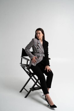 Şık genç iş kadını bir sandalyede oturuyor ve başka tarafa bakıyor, modern bir ceket ve topuklu ayakkabı giyiyor, özgüveni, bağımsızlığı ve profesyonel modayı simgeliyor..