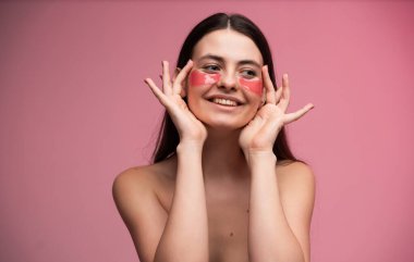 Genç bir kadın, temiz ciltli, doğal makyajlı ve modern cilt bakımlı güzel stüdyo fotoğraflarına pembe hidrojel göz bantları yapıştırıyor.