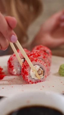 Bir kadın elinin beyaz bir tabaktan suşi rulosunu alıp soya sosuna batırması ve taze deniz ürünleriyle geleneksel Japon mutfağının tadını çıkarması.