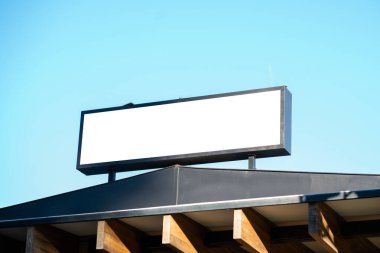 Ticari bir binanın çatısında bulunan boş beyaz reklam, pazarlama kampanyaları veya açık mavi gökyüzü altında marka tanıtımı için temiz bir şablon sunuyor.