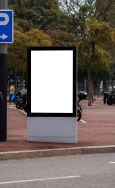 Boş dikey sokak reklam panosu modeli, özelleştirilebilir beyaz bir ekran sağlıyor, motosiklet park alanı ve arka planda yemyeşil ağaçlar bulunan kentsel bir ortamda.
