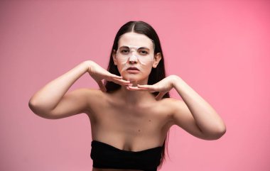 Hidratlaştırıcı maske takan, elleri çenesinin altında poz veren, güzellik ve cilt bakımını teşvik eden genç bir kadın hayat dolu pembe bir arka plana karşı kendini korumak için.