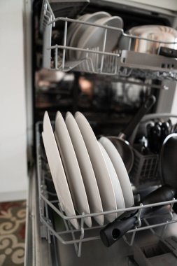 Bulaşık makinesinde yeni temizlenmiş beyaz tabaklar, tencereler ve çatal bıçaklarla dolu raflar, modern ev verimliliğini ve günlük rutinini temsil ediyor.