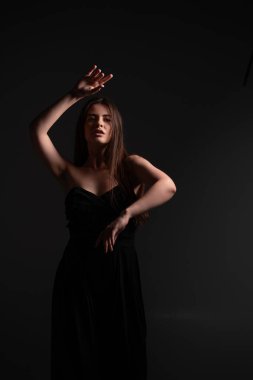 Karanlık bir stüdyoda duran, zarif siyah bir elbise giyen, etkileyici el hareketleriyle poz veren, ciddi bakışları olan, sanatsal bir hareketi ve modayı yakalayan bir kadın.