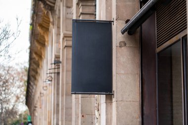 Eski bir tarihi binanın duvarına asılmış boş siyah dikey tabela, şehrin arka planındaki markalaşma reklamı için makara, metin için metin alanı