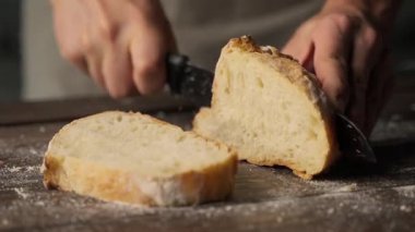 Fırıncının elleri, un tozlu ahşap bir masada taze, kabuklu bir ekmek dilimlemek için bıçak kullanırken.