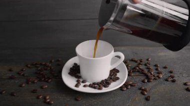 Beyaz seramik bir fincana Fransız presinden sıcak sade kahve döken biri. Kavrulmuş kahve çekirdekleri koyu ahşap masaya saçılmış.