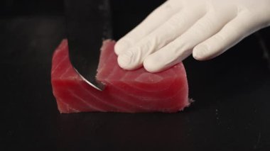 Profesyonel suşi şefi beyaz eldivenler giyip siyah bir tahtanın üzerinde taze çiğ ton balığını dilimliyor ve yüksek kaliteli balıkları geleneksel bir Japon yemeği için hazırlıyor.