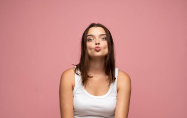 Genç esmer kadın komik bir öpücük yüzü yapıyor, pembe arka planda neşeli bir görünüm için dudaklarını büküyor.