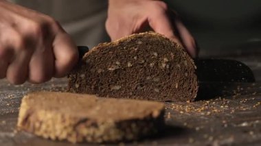 Fırıncının elleri, dişli bir bıçakla, taze, karanlık, tam tahıllı bir ekmeği, ahşap bir yüzeyde dilimlemek için yakın plan çekiliyor.