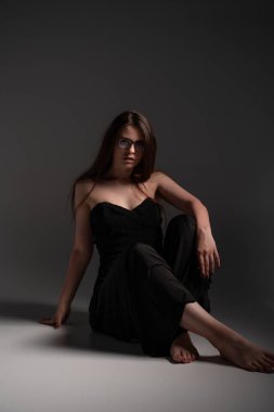 Genç bir kadın, siyah askısız elbise içinde, gözlük takıyor, zarafet, zeka ve modern moda bir stüdyo resminde kendine güvenerek poz veriyor.