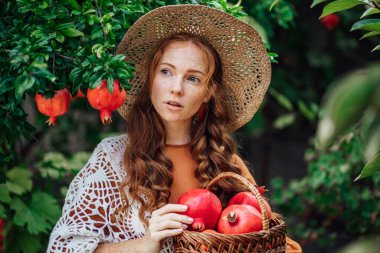 Kırmızı nar bahçesi, turuncu elbiseli ve beyaz şallı mutlu kızıl saçlı kız. Büyük kırmızı meyveli kadın portresi
